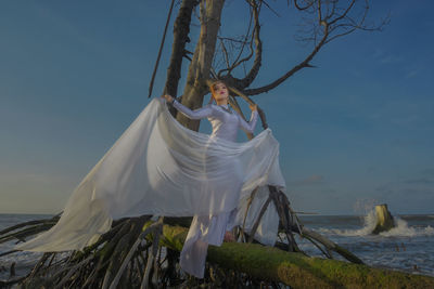 Low angle view of young woman with scarf posing on bare tree at beach