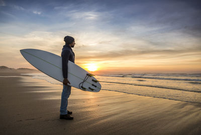 Man with surfboard on beach against sky during sunset