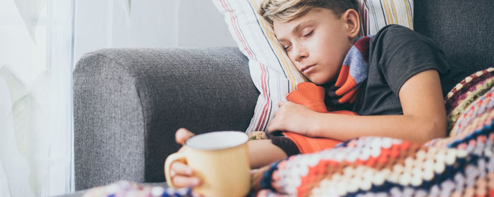 Boy sleeping on sofa with blanket while holding coffee cup in hand at home