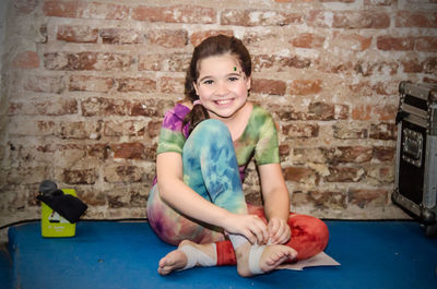 Portrait smiling girl sitting on floor against brick wall