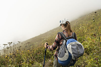 Young woman in plaid shirt with backpack trekking poles walking along mountain trail hiking in fog