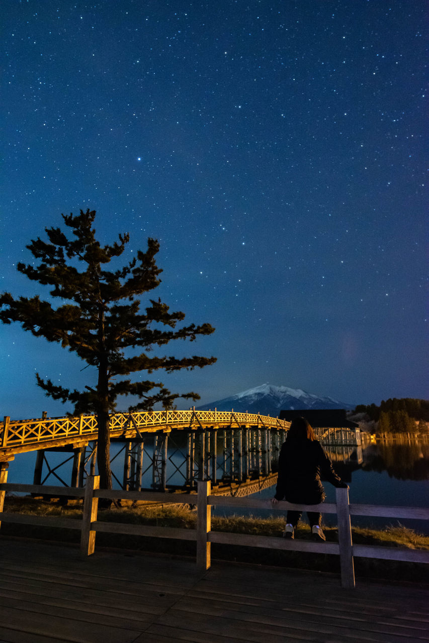 REAR VIEW OF MAN SITTING AT RIVERBANK AGAINST SKY