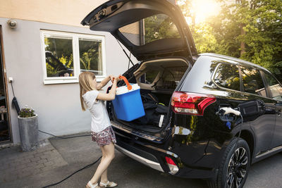 Girl loading cooler in black car trunk against house