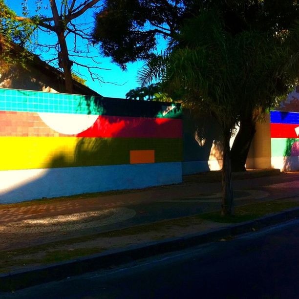 architecture, built structure, building exterior, tree, transportation, street, road, sunlight, graffiti, multi colored, city, sky, outdoors, day, car, house, road sign, text, no people, shadow