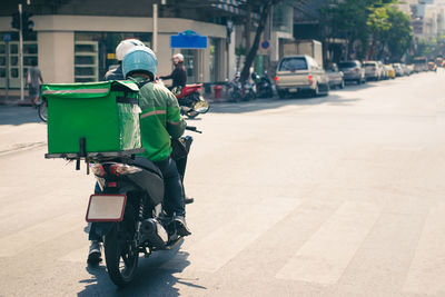 Rear view of delivery person riding motorcycle on road in city