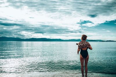 Rear view of woman standing in swimwear at lakeshore against cloudy sky