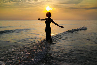 Silhouette woman with arms outstretched standing in sea against orange sky