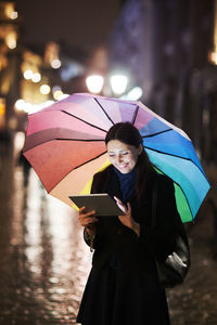 Smiling woman with umbrella using digital tablet while standing on wet road at night