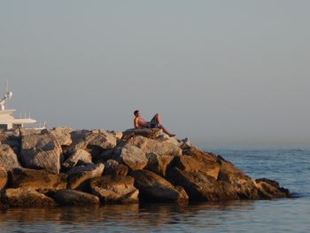 Shirtless man relaxing on rocks by sea against sky
