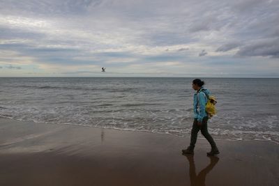 Side view full length of woman walking on shore against cloudy sky