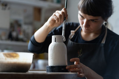 Happy concentrated young craftswoman paints clay vase sitting at work table closeup