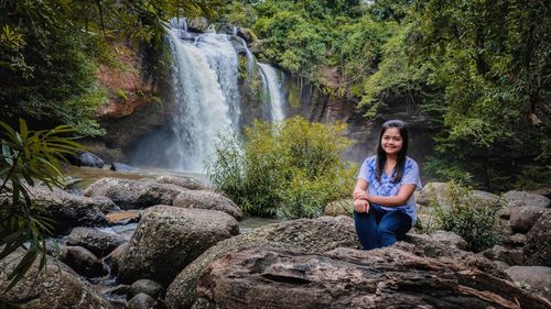 Portrait of woman sitting on rock against waterfall in forest