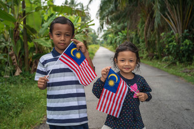 Portrait of siblings holding malaysian flag while standing on road amidst plants