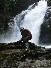 Man crouching on rock against waterfall