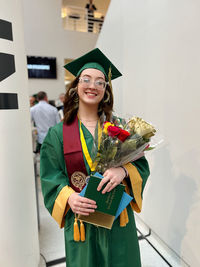 Portrait of young woman wearing graduation gown standing against wall