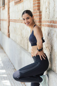 Smiling fit sportswoman in leggings and bra leaning brick building and looking at camera while relaxing after training