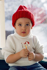 Boy in white sweater and red knitted hat sits on the windowsill and eats gingerbread horse on stick