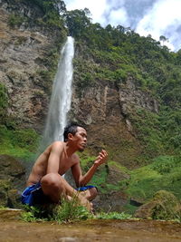 Full length of shirtless man on rocks against waterfall