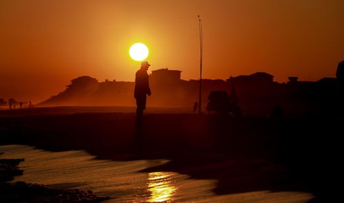 Silhouette people standing on shore against orange sky