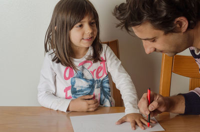 Father assisting daughter while drawing on white paper against wall