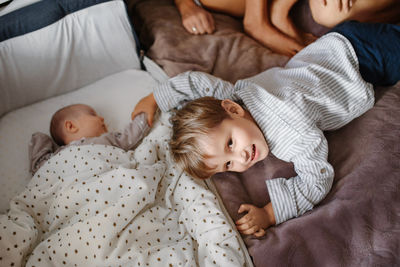 Toddler boy lies on the bed next to a bedside bassinet and gently touches the newborn hand