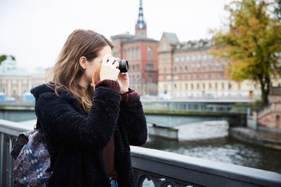 Young woman photographing through camera while standing on bridge in city