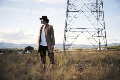 Contemplative young hipster male in stylish coat and hat standing in grassy field near metal electric tower and looking away thoughtfully while spending autumn day in countryside