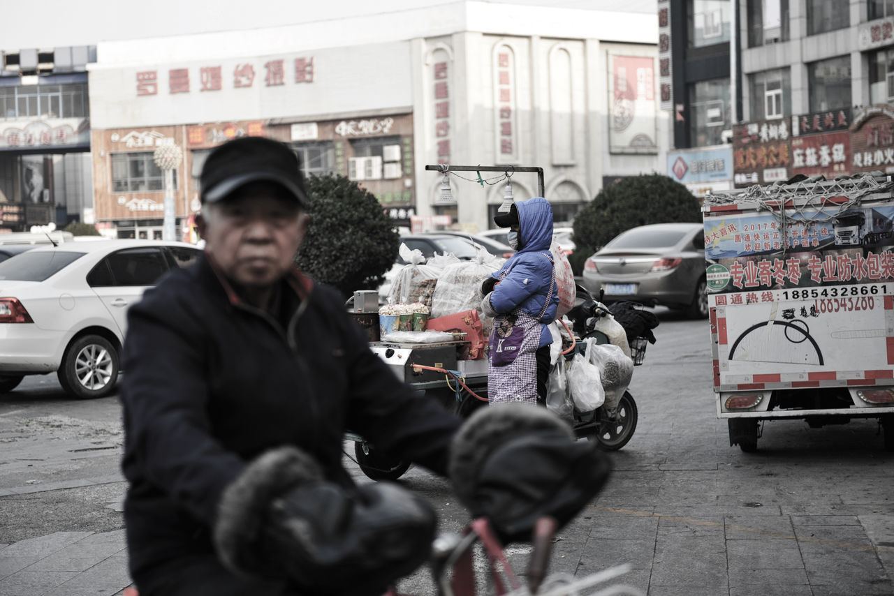 mode of transportation, city, motor vehicle, car, transportation, building exterior, land vehicle, architecture, street, one person, real people, standing, built structure, men, day, lifestyles, incidental people, adult, city life, city street, outdoors, warm clothing