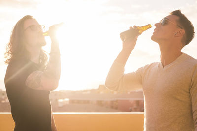 Male friends drinking beer while standing against sky during sunset