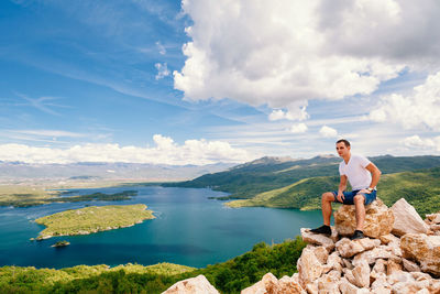 Man sitting on rock at lake against sky