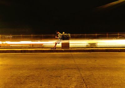 Man using mobile phone while sitting by light trails on road at night