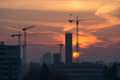 Silhouette crane by building against sky during sunset