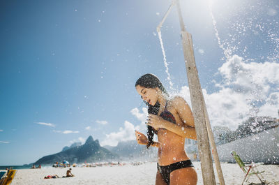 Woman bathing below shower at beach against sky