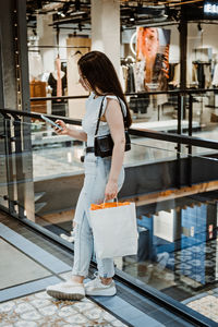 Young latina woman with shopping bags using her smart phone while standing in shopping center, mall