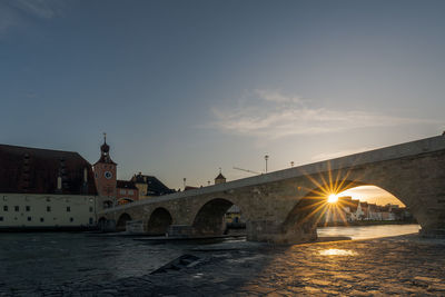 Arch bridge over river by buildings against sky during sunset