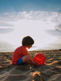  boy sitting at the beach with a red ball with a sea at the background 