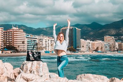 Full length of young woman standing on rock by sea against sky