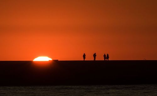 Silhouette people standing by sea against orange sky