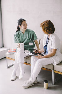 Full length of female doctor holding coffee cup while discussing with nurse sitting on sofa at hospital