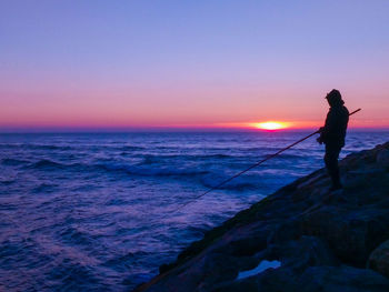 Silhouette man fishing in sea against sky during sunset