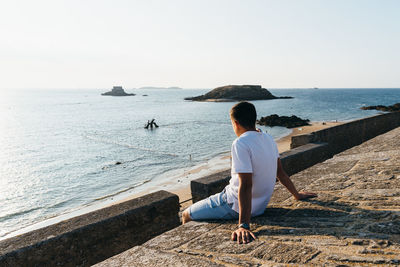 Side view of teenage boy looking at sea while sitting on retaining wall against clear sky