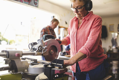 Senior woman using circular saw while man standing in background at workshop