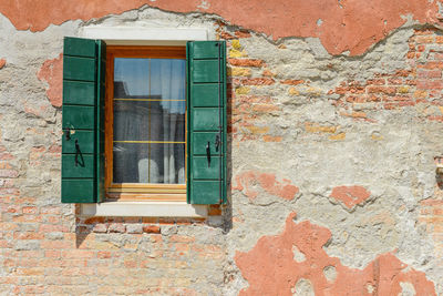 Green window on orange old house wall cracks in concrete visible red bricks in burano, italy.