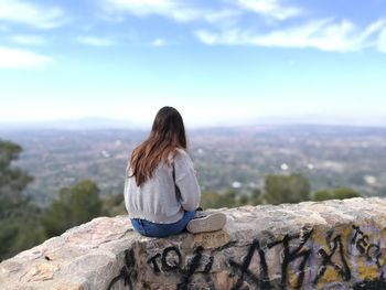 Rear view of woman sitting on retaining wall against sky