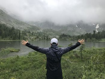 Rear view of man with arms outstretched standing by plants against lake and mountain