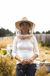 Portrait of beekeeper wearing protective mask and hat while standing on field against clear sky