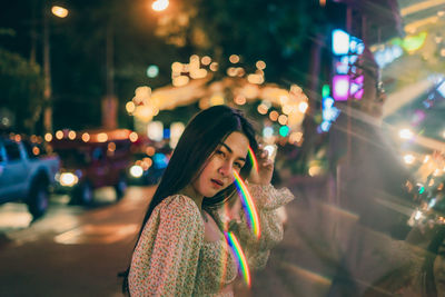 Portrait of young woman standing on illuminated street in city at night