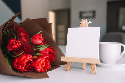 A bouquet of red tulips on the table and a white cup of coffee, empty white frame. greeting card