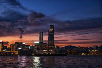 Illuminated modern buildings by river against sky at sunset