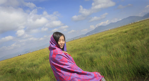 Portrait of woman wrapped in shawl sitting by grassy landscape against sky